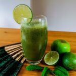 A tall glass of cucumber apple smoothie, surrounded with an apple, limes, and mint leaves, with a hand fan lying next to the glass