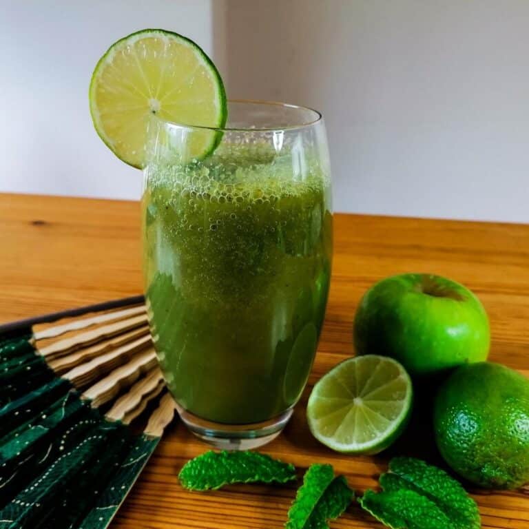 A tall glass of cucumber apple smoothie, surrounded with an apple, limes, and mint leaves, with a hand fan lying next to the glass