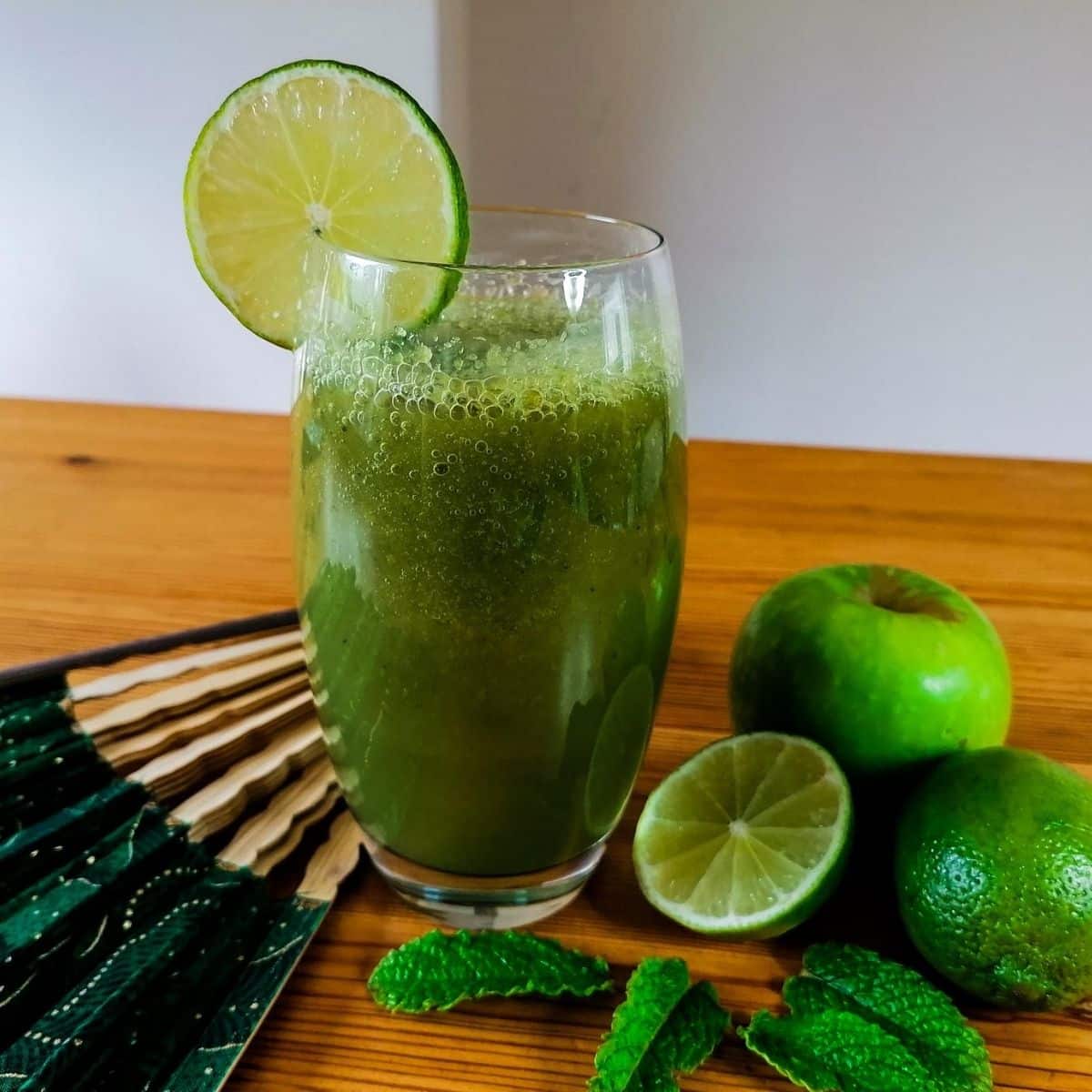 A tall glass of cucumber apple smoothie, surrounded with an apple, limes, and mint leaves, with a hand fan lying next to the glass