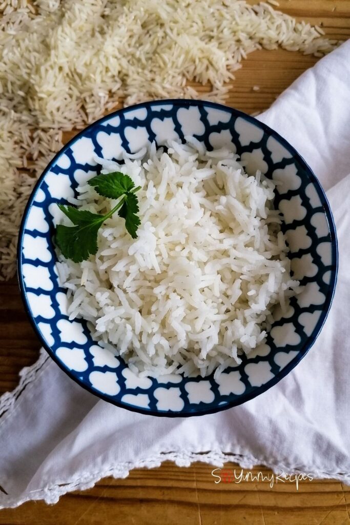 A blue patterned bowl of cooked basmati rice garnished with coriander leaves. Underneath there is a white napkin and some rice grains