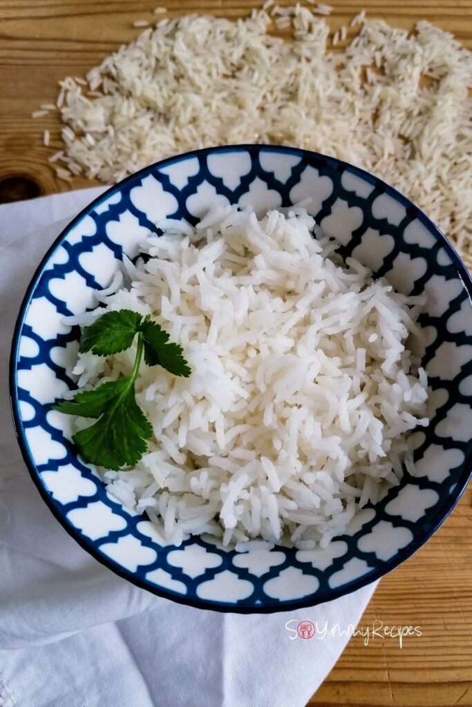 cooked basmati rice garnished with coriander leaves in a blue patterned bowl with a white napkin underneath the bowl and some sprinkles of rice grains around