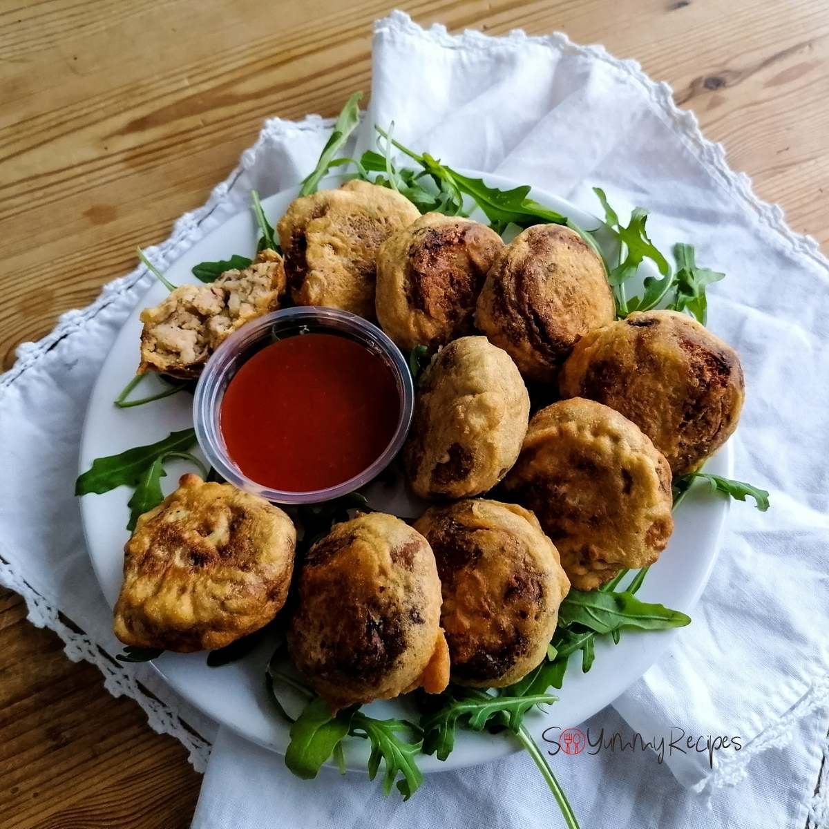 a plate of Pakistani Aloo Tikki on a white napkin with a small bowl of chilli sauce and some rocket leaves.