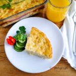 a slice of macaroni schotel with slices of cucumber and a plum tomato on a white plate with a glass of orange juice next to it