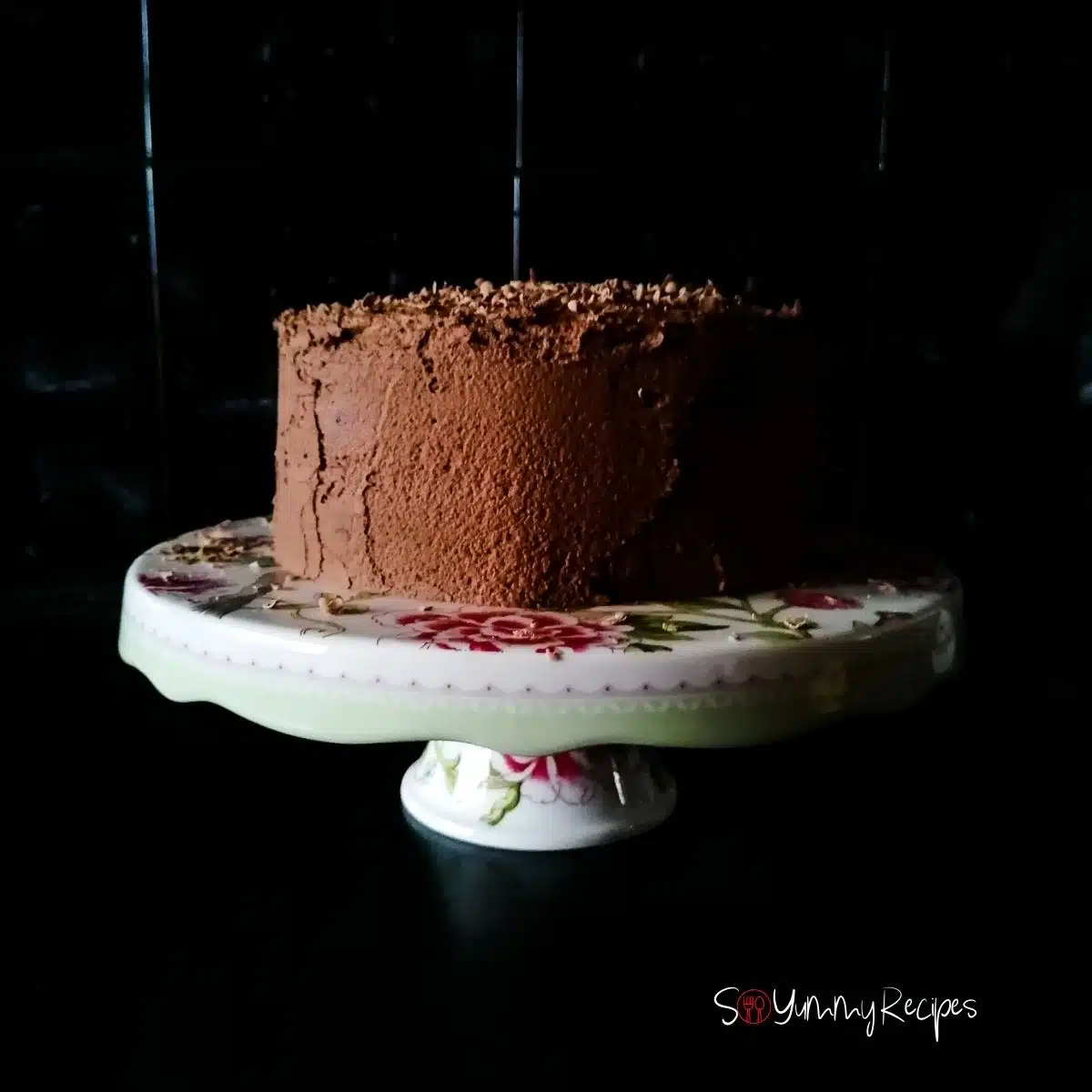 chocolate cake with chocolate mousse frosting on a white cake stand with black tile background