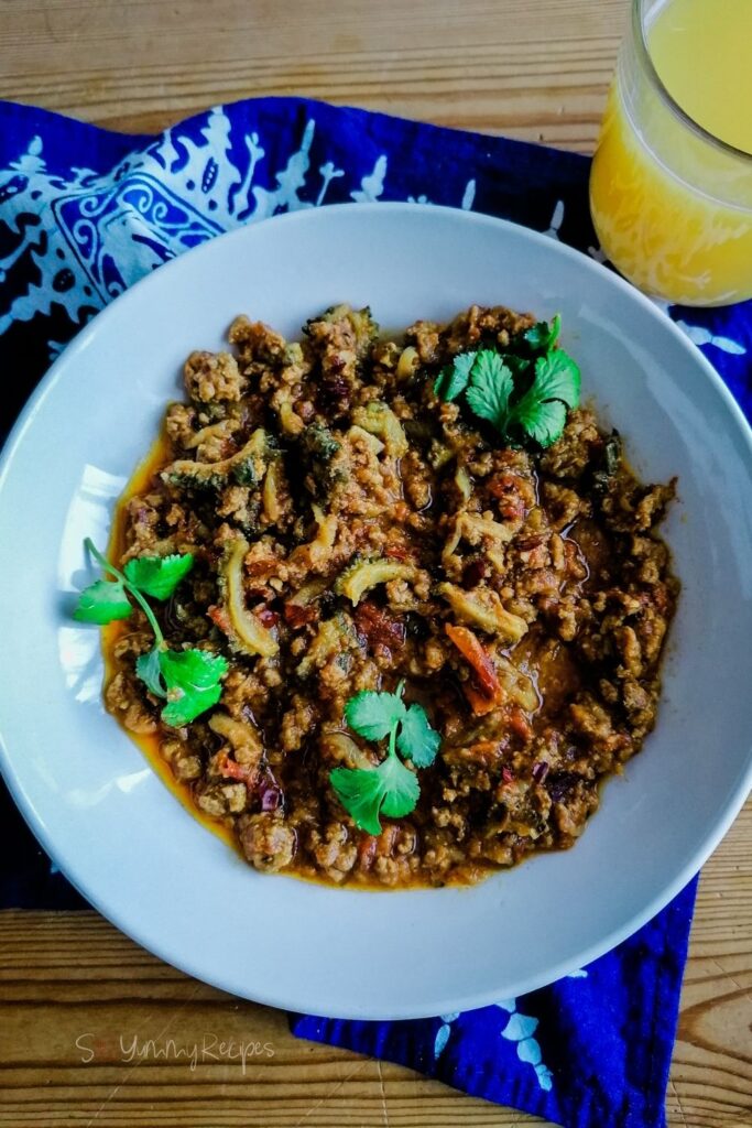 a plate of minced meat curry with bitter gourd and a glass of orange juice on the side with blue patterned napkin underneath the plate