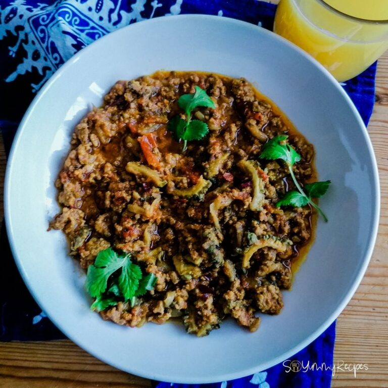A dish of Keema Karela - the minced meat curry with bitter gourd with blue patterned napkin underneath the dish and a glass of orange juice on the side of the dish