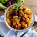 Fruit chaat in Pakistani style in a crystal bowl with a white napkin, a spoon and some fruits in the background
