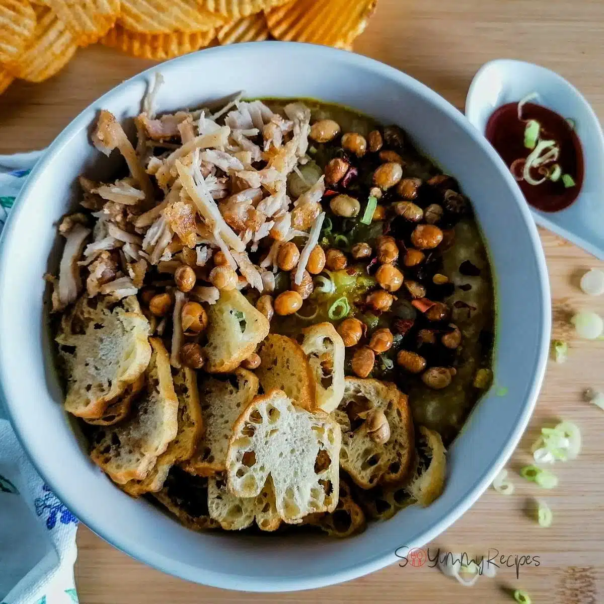 A bowl of Indonesian rice porridge - Bubur Ayam - topped with shredded chicken, cakwe, fried soya beans, with a spoon of soy sauce and, potato crisps, and spring onion slices on the side.