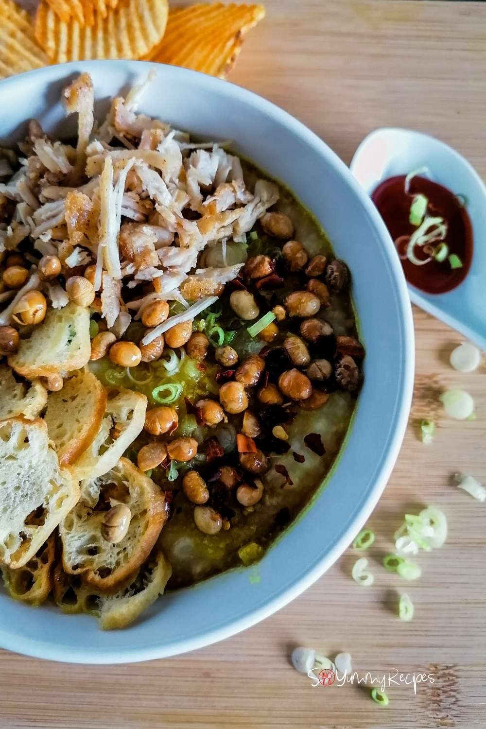 A bowl of Bubur. Ayam - the Indonesian rice congee with shredded chicken, fried soya beans, and cakwe as garnishes with a spoon of soy sauce and spring onion slices on the side.