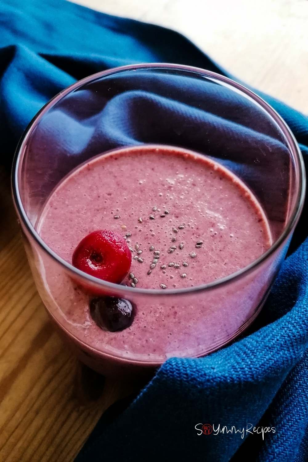 a glass of cherry smoothie with cherries on the top and blue napkin around the glass.
