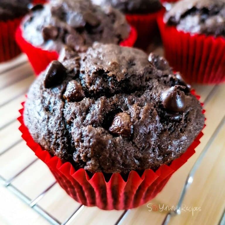 A double chocolate chip muffin on a red muffin case on a cooling rack with a few other muffins in the background.