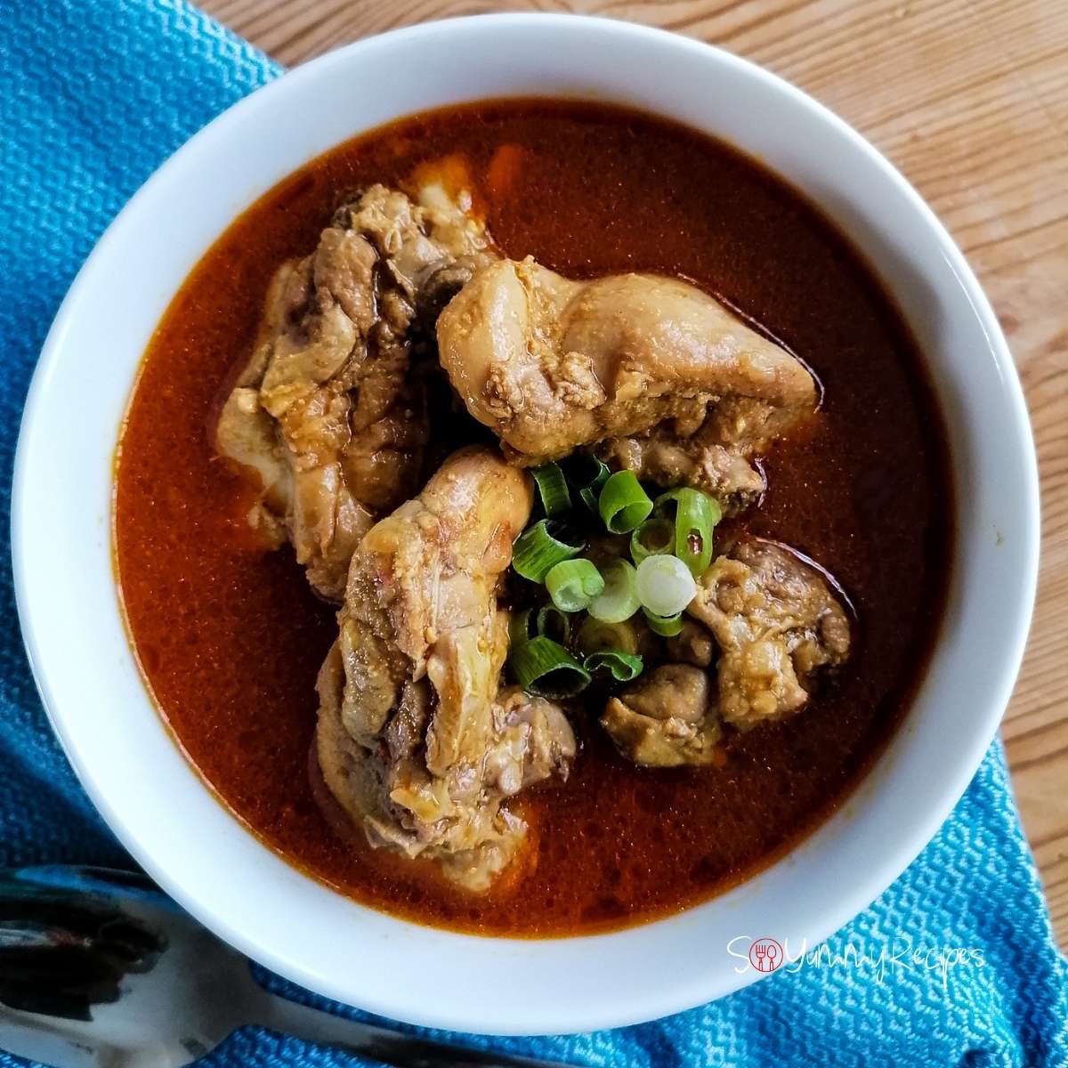 A white bowl of Indonesian chicken curry - Kari Ayam - on wooded table with blue tea towel on the left side.