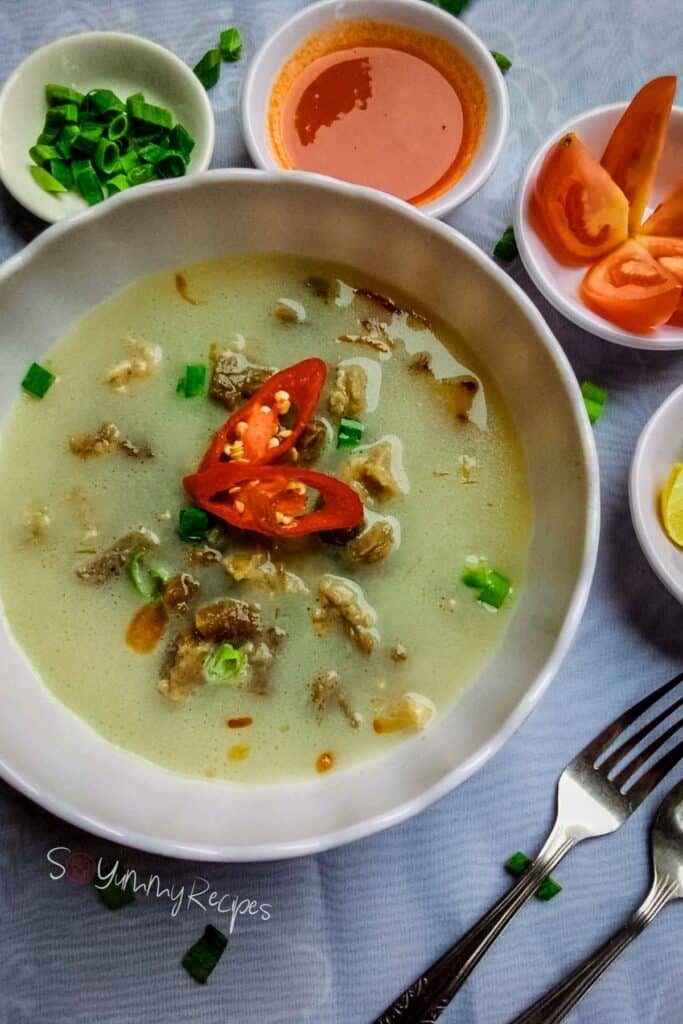 Indonesian beef soup from Jakarta in a round bowl with some condiment, tomatoes, spring onions and lemon in small bowls.