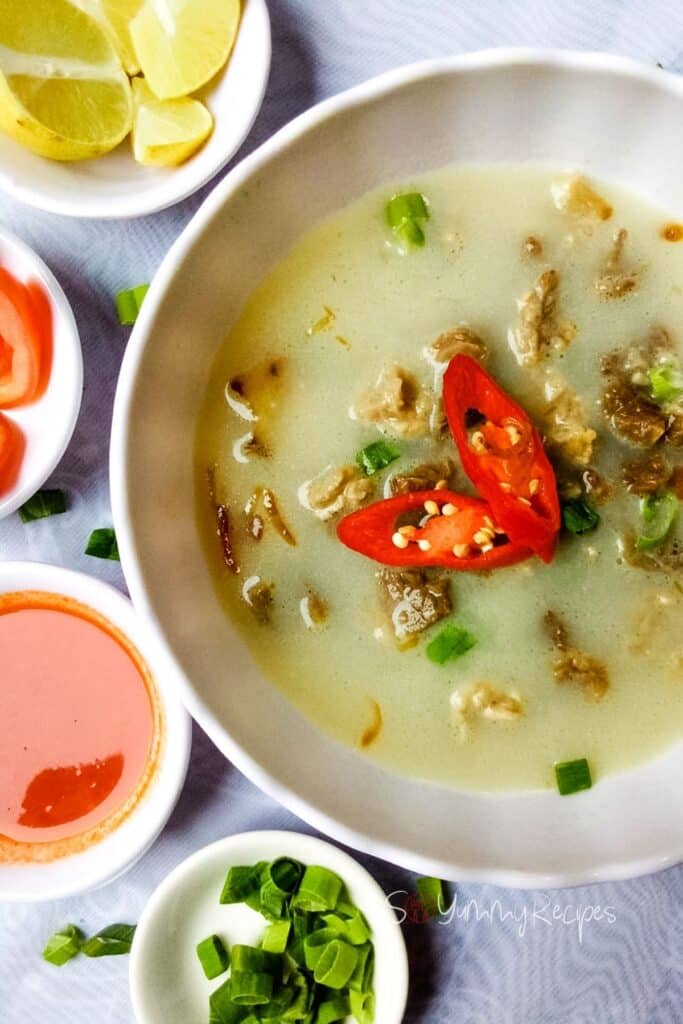 Soto Betawi the beef soup from Jakarta region in a white round bowl surrounded with condiments, lemon, tomato and spring onion in small bowls.