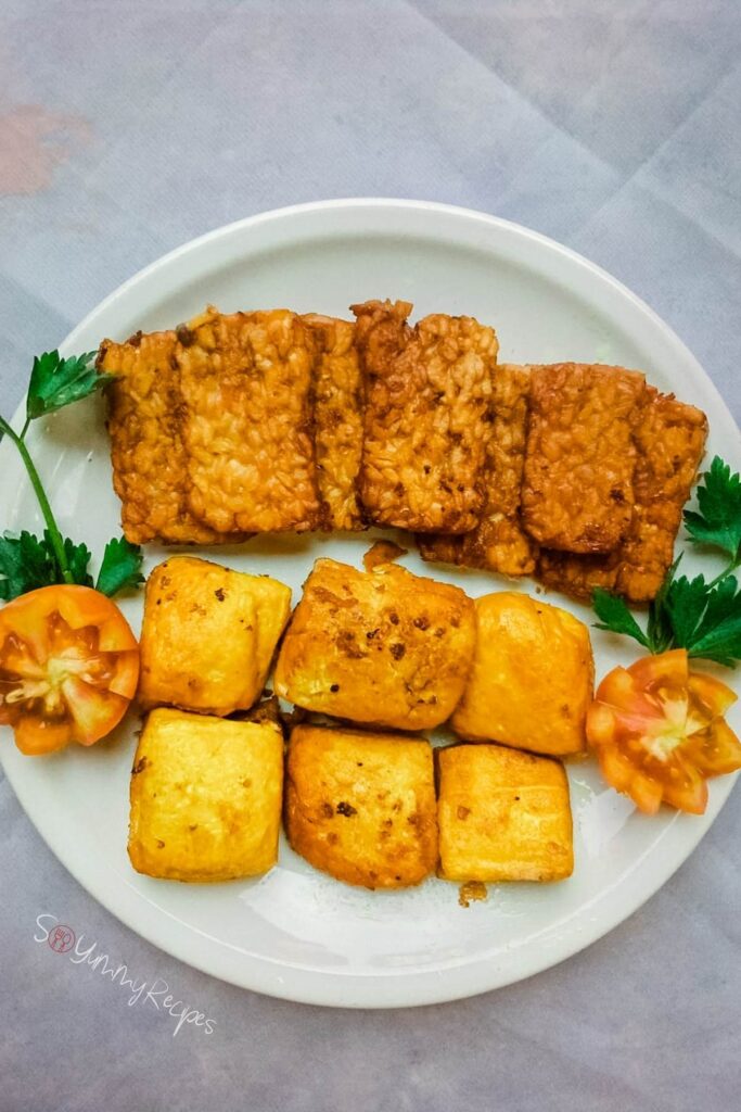 A round dish full of tahu and tempe bacem wih parsley leaves and tomato as garnish.