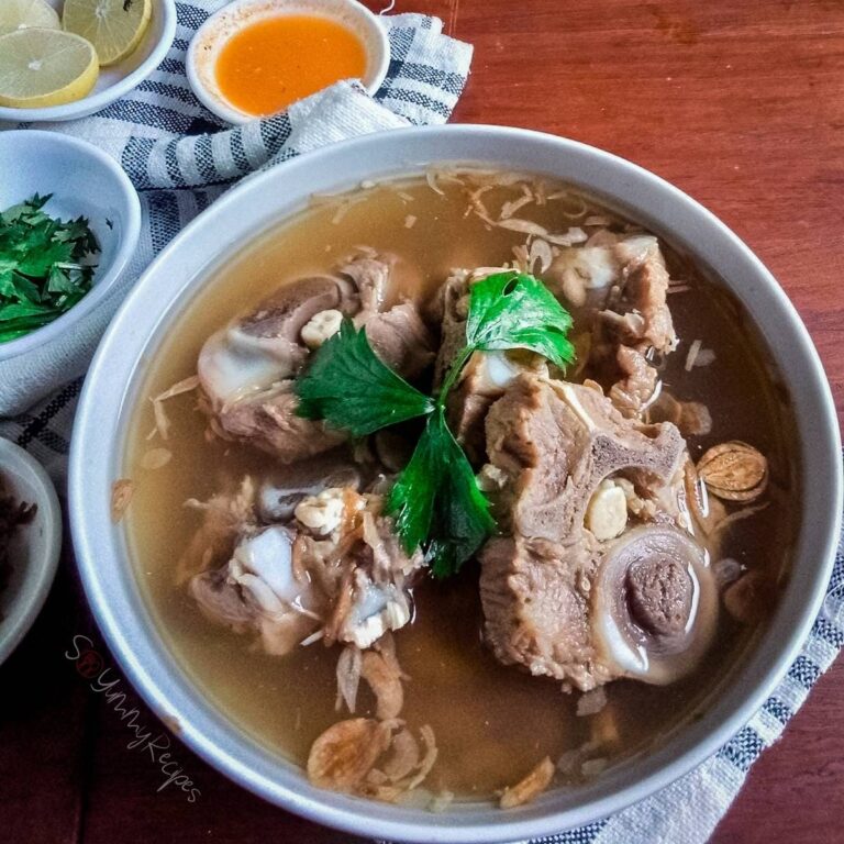 A bowl of Indonesian oxtail soup which is called Sop buntut with small bowls around it containing lemon slices, chilli sauce, chopped parsley.