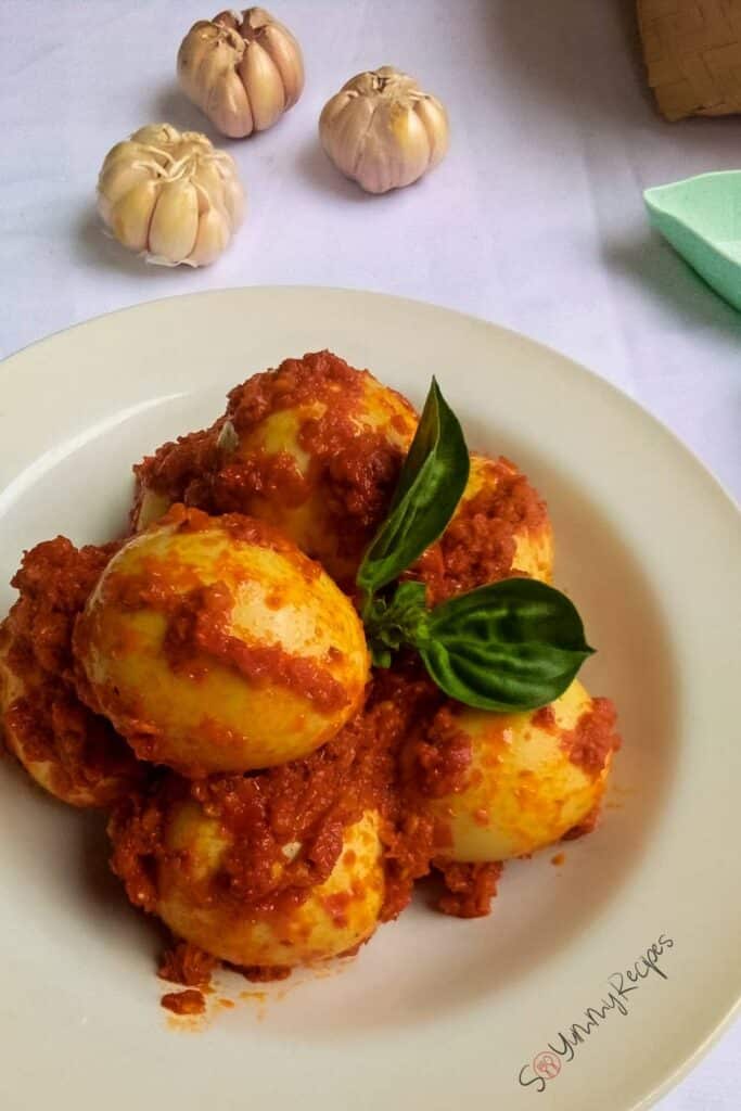 Telur Balado the Indonesian hard boiled eggs in spicy chilli sambal on a white dish with three heads of garlic on the background.
