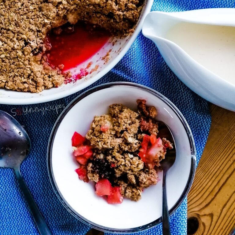 Apple blackberry crumble in a serving bowl with a jar of custard and the crumble dish next to it.