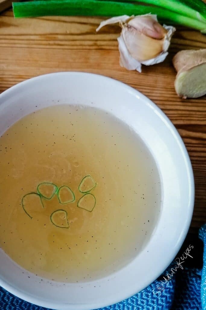 Clear chicken stock in white bowl with garlic and spring onion on the background.