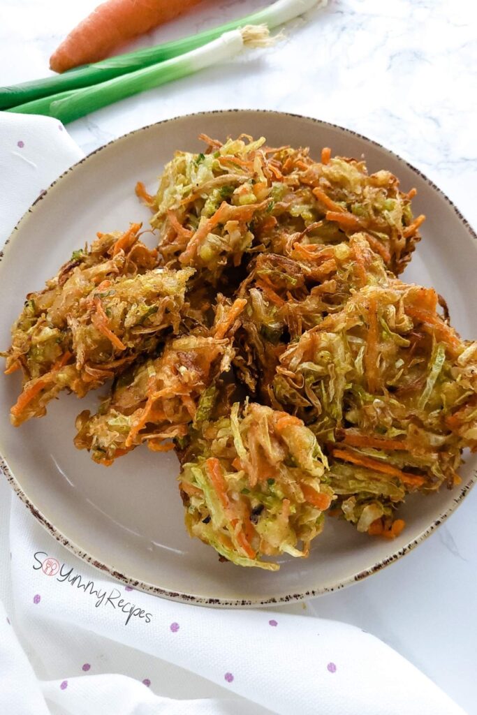 Vegetable fritters on a round plate with spring onion and carrot in the background.