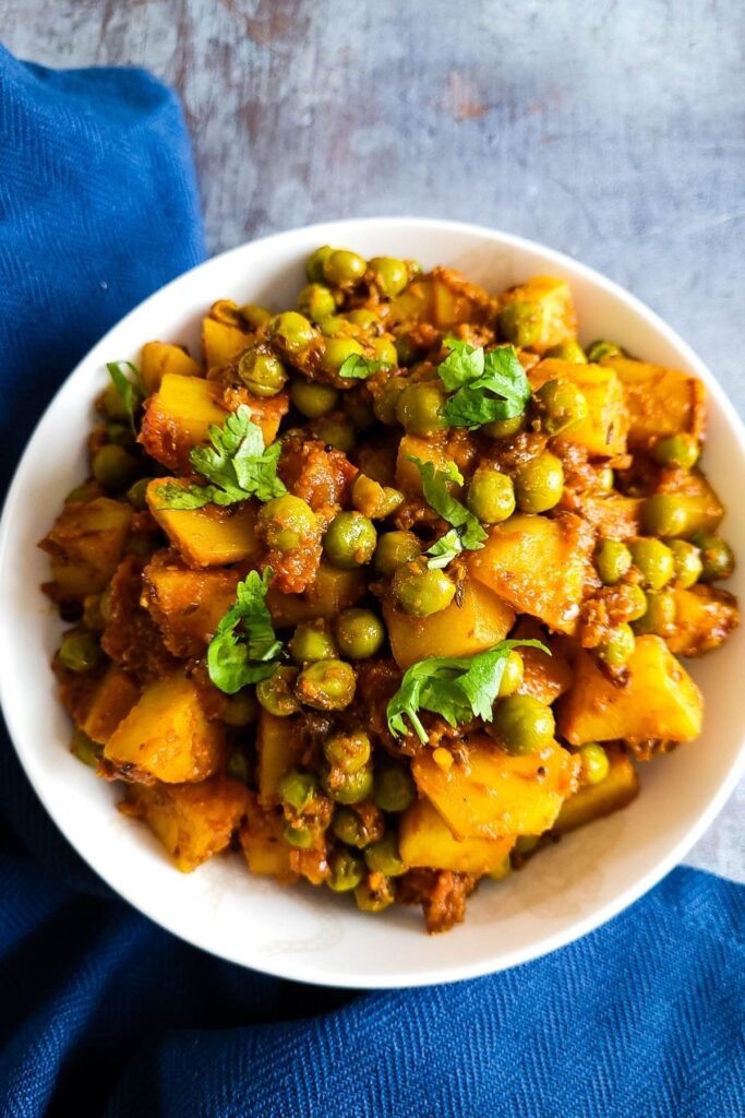 Aloo Matar - Pakistani potato and green pea curry in a white bowl with blue napkin on the background.