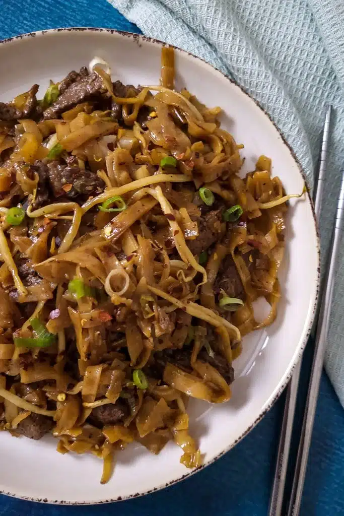 Chinese stir-fry flat rice noodles Beef Ho Fun on a white plate with a pair of metal chopsticks next to it.