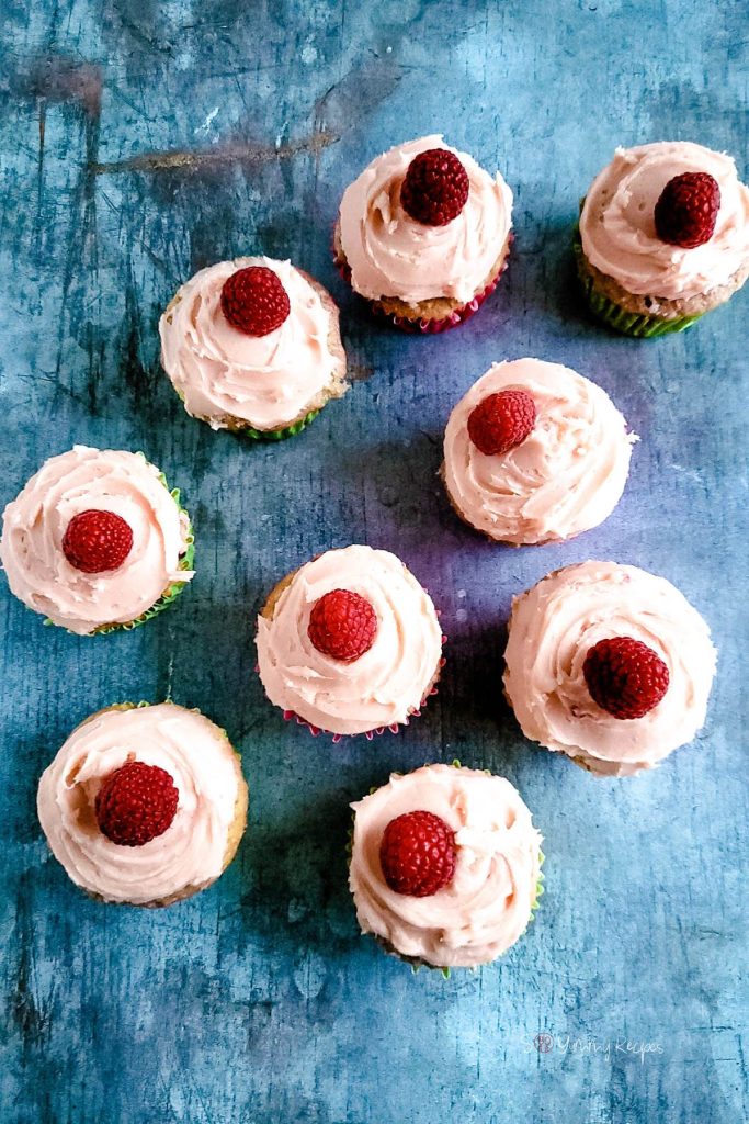 Nine cupcakes with pink buttercream and topped with raspberries.