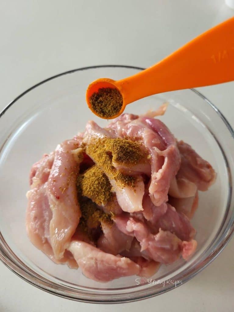 Chicken pieces in a bowl and a spoon of ground cumin over it.