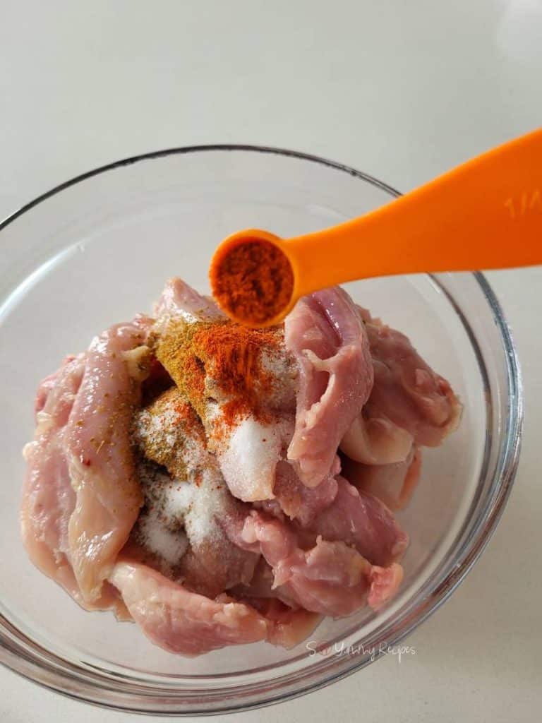 A spoon of red chilli pepper powder over a bowl of chicken pieces.