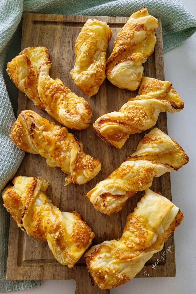 Puff pastry cheese straws on a wooden chopping board with a mint green kitchen towel next to it.