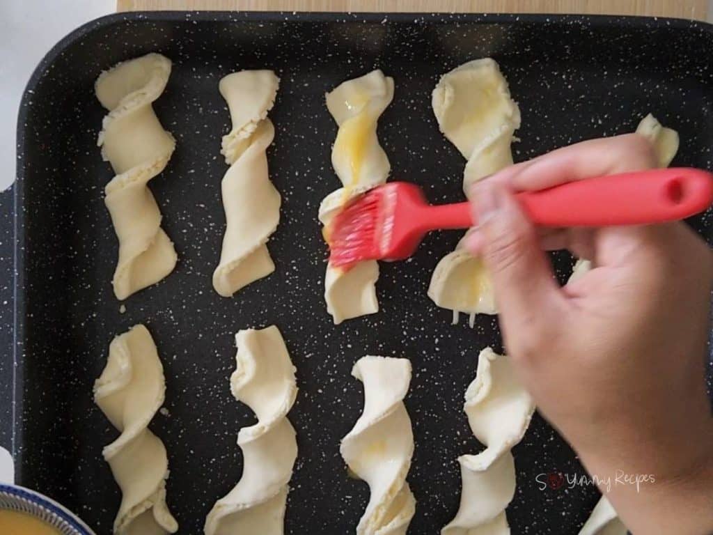 A hand holding pastry brush over pastry straws that lined on a baking tray.