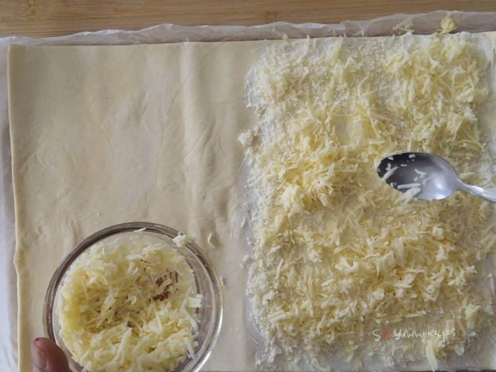 Ready-rolled puff pastry with cream cheese and grated cheese on the top, a bowl of grated cheese and a teaspoon.