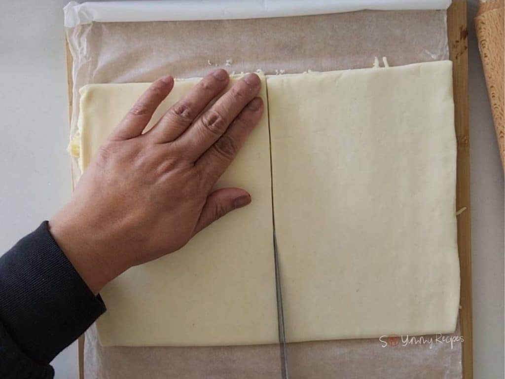 A hand over a puff pastry with a knife cutting halfway.