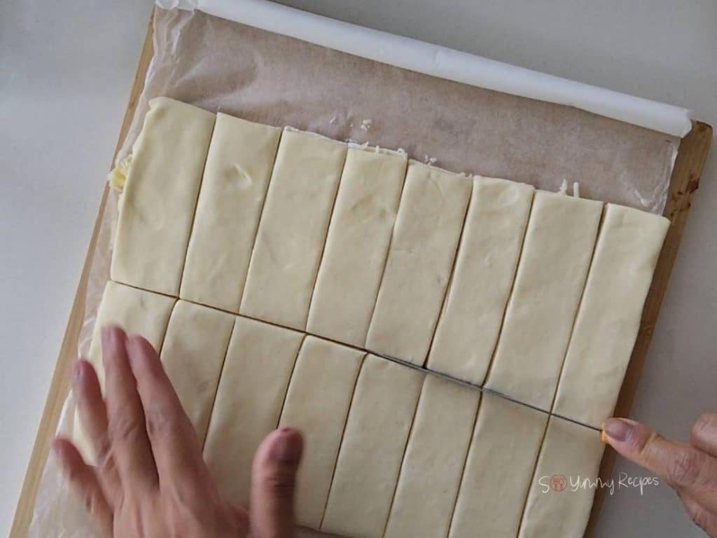 A hand holding a puff pastry strips with a knife cutting it.