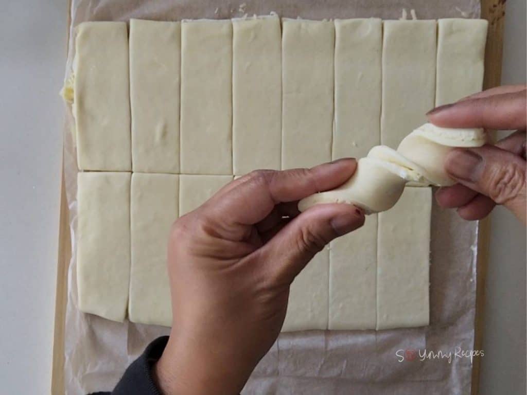 Hands twisting a strip of puff pastry and a sheet of puff pastry in the background.