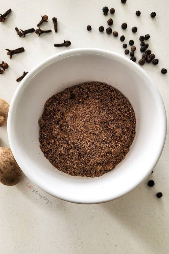 Garam masala spice mix in a white bowl with whole spices in the background.