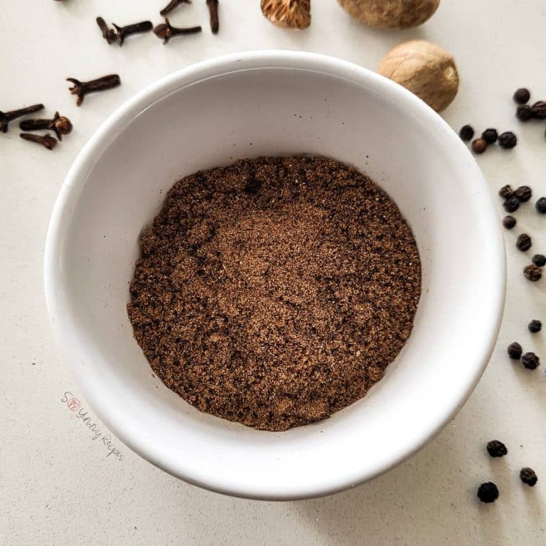 Garam masala in a small white bowl surrounded with whole spices.