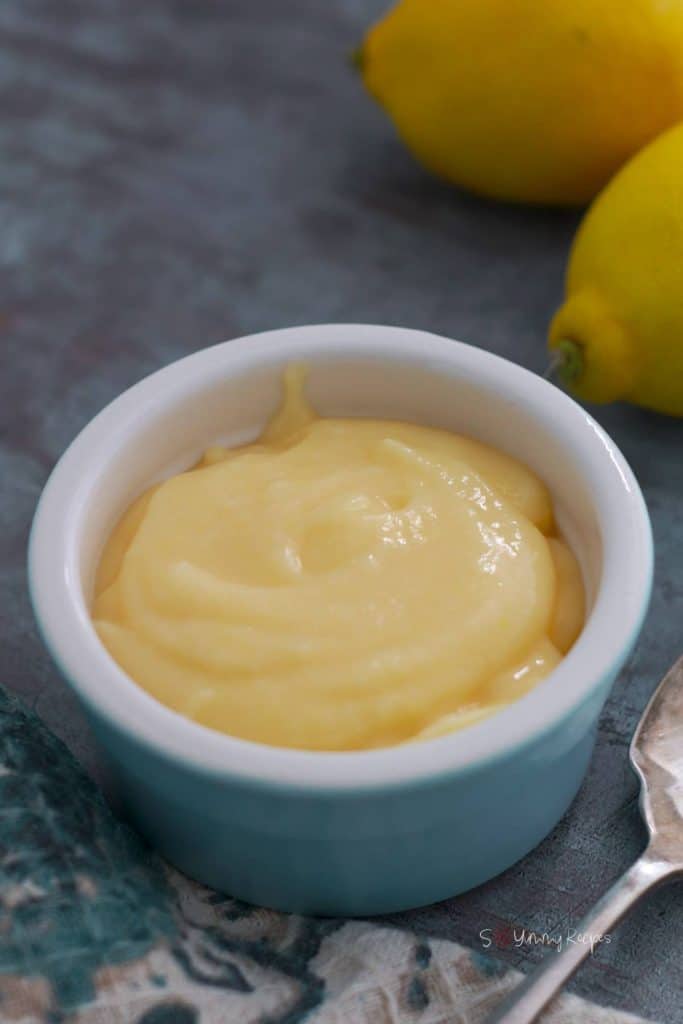 A small bowl of lemon curd with a few lemons in the background.