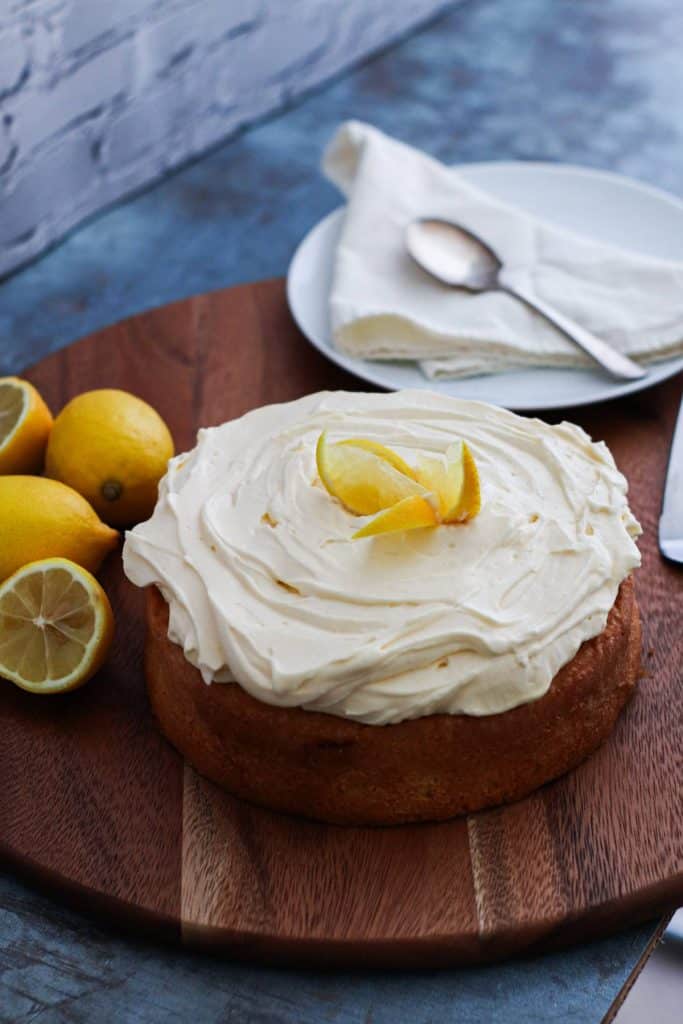 A lemon cake frosted with cream on a wooden chopping board with lemons, a plate, a spoon on the background.