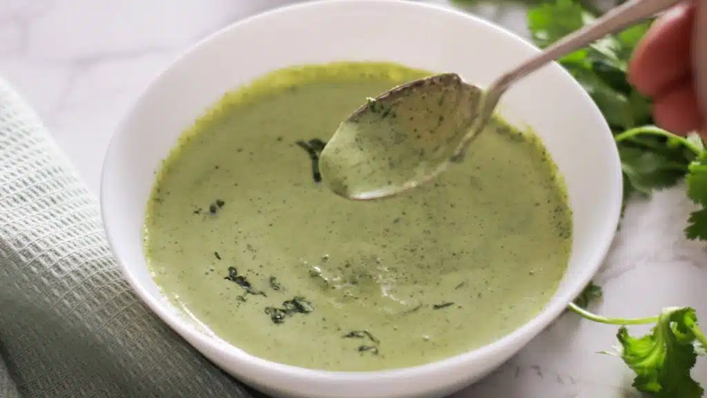 Green coriander and mint chutney in a white bowl with spoon over it.