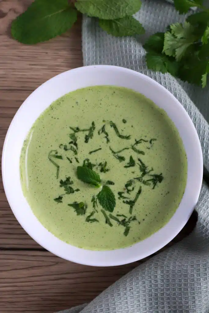 A bowl of green chutney with green napkin, mint and cilantro in the background.