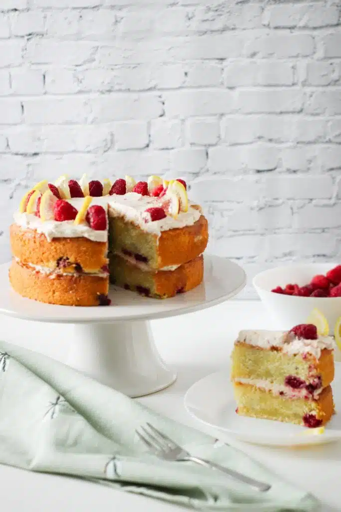 Raspberry lemon cake on a cake stand with a slice of it on a cake plate next to it.