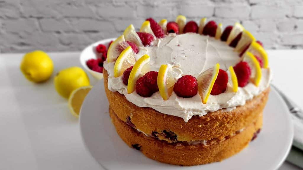 Lemon raspberry cake on a white cake stand with lemons in the background.