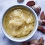 Garlic paste in a white bowl with some garlic cloves in the background.