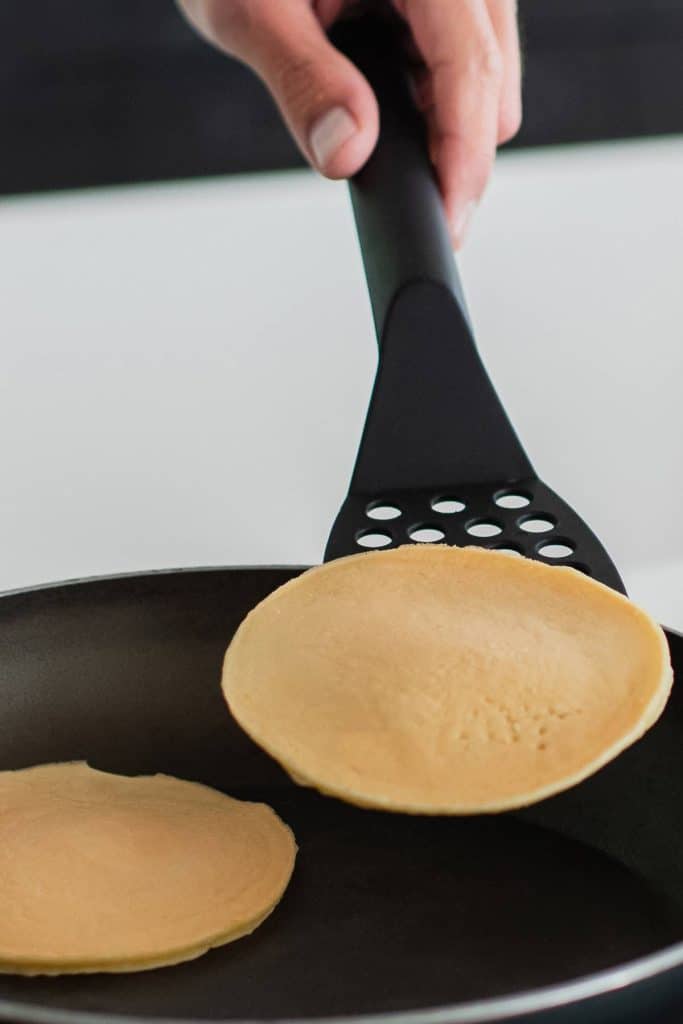 A hand holding a slotted spoon with a pancake on it and another pancake on a frying pan.