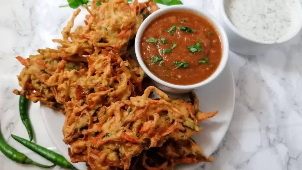Vegetable pakoras with a bowl of tomato chutney and mint yogurt sauce with some green chilies in the background.