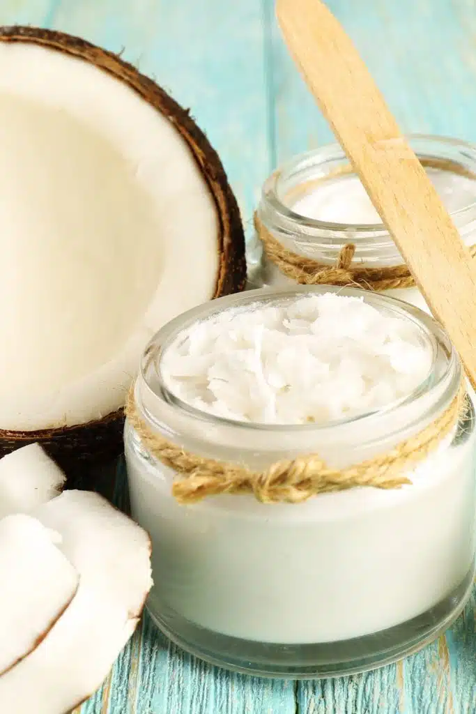 A jar of coconut oil with raw coconuts and wooden spoon nearby.