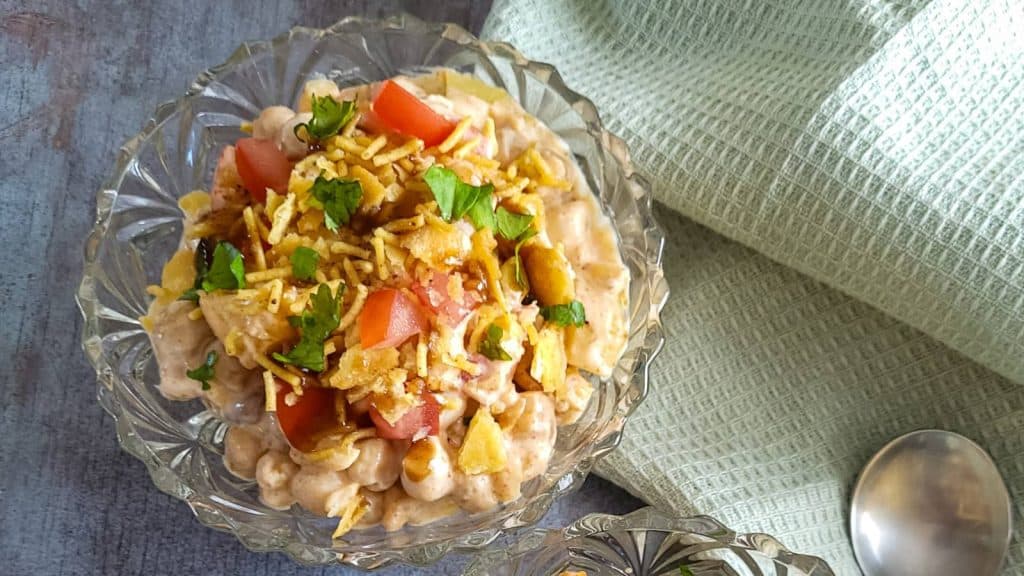 A bowl of Papdi chaat with mint green napkin and a spoon on the side.