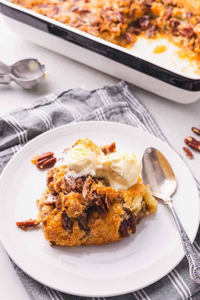 A portion of pecan cobbler on a white plate with a spoon and the dish next to it.