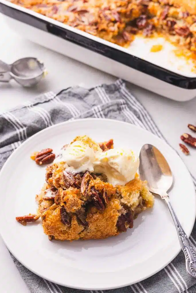 A portion of pecan cobbler on a white plate with a spoon and the dish next to it.